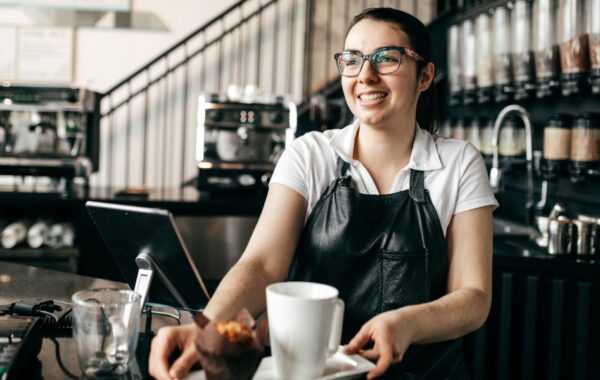 Photo of a barista wearing an apron. They are behind counter in coffee shop. They are smiling as they pass a tray over the counter which has a cup and muffin on it.