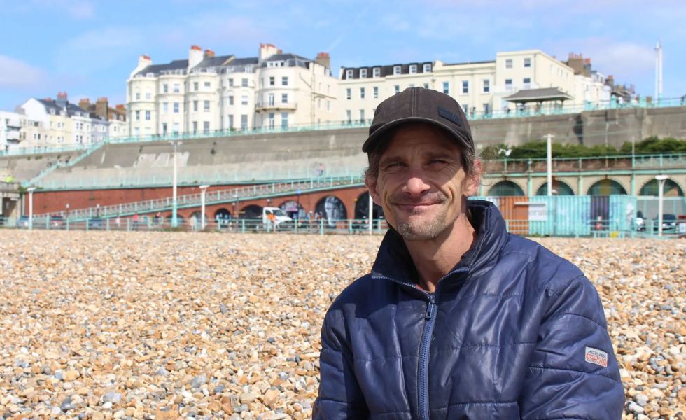 Close up photo of someone sitting on beach smiling to camera with Kemptown seafront buildings in the background.