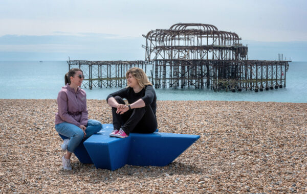 Two women are sat on a star shaped monument on Brighton beach, with the West Pier behind them.