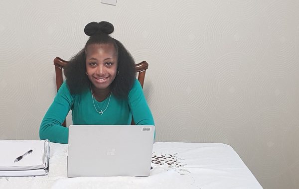 A black woman wearing a green top sits at her laptop smiling at the camera