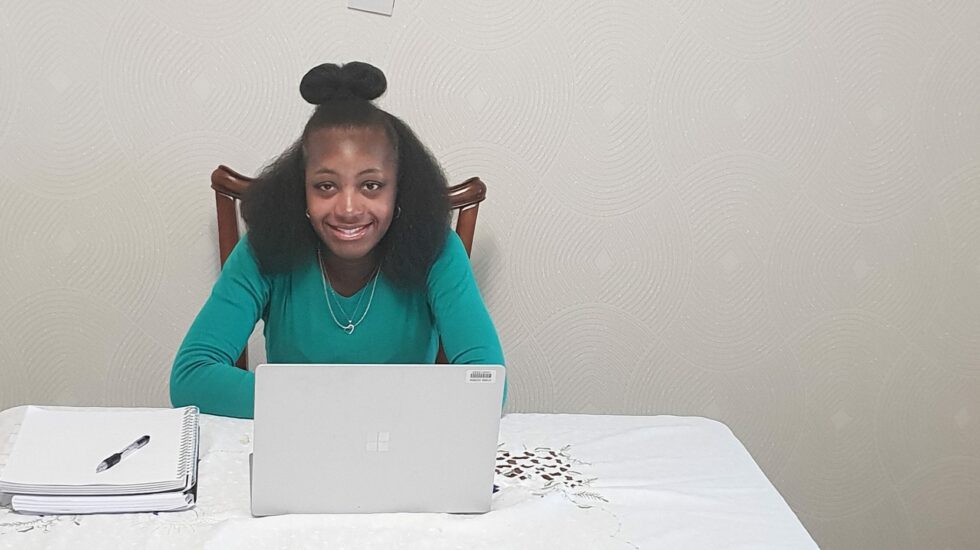 A black woman wearing a green top sits at her laptop smiling at the camera