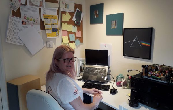 A woman with long blonde hair, wearing glasses and a white t-shirt sits at her home desk