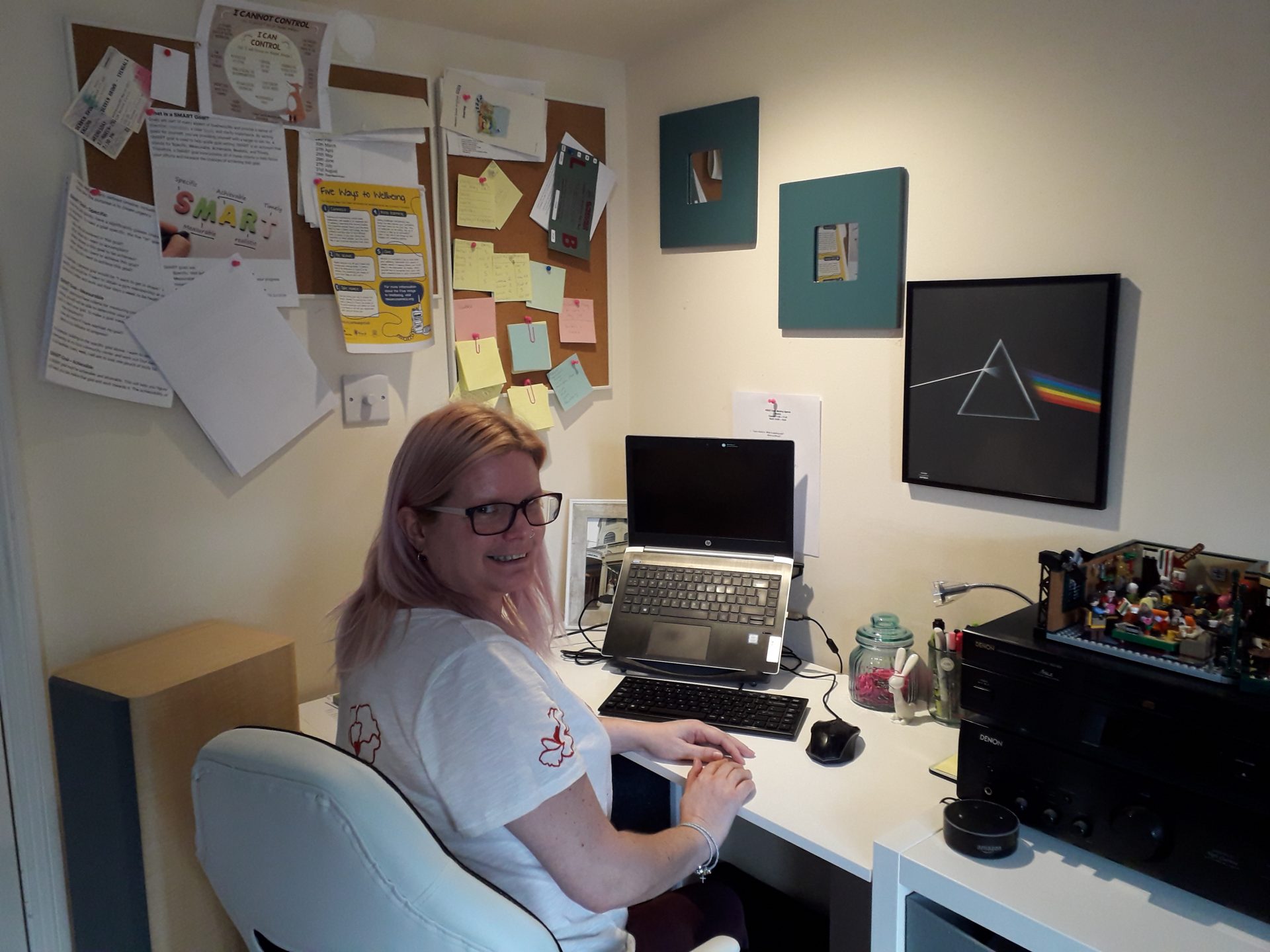 A woman with long blonde hair, wearing glasses and a white t-shirt sits at her home desk