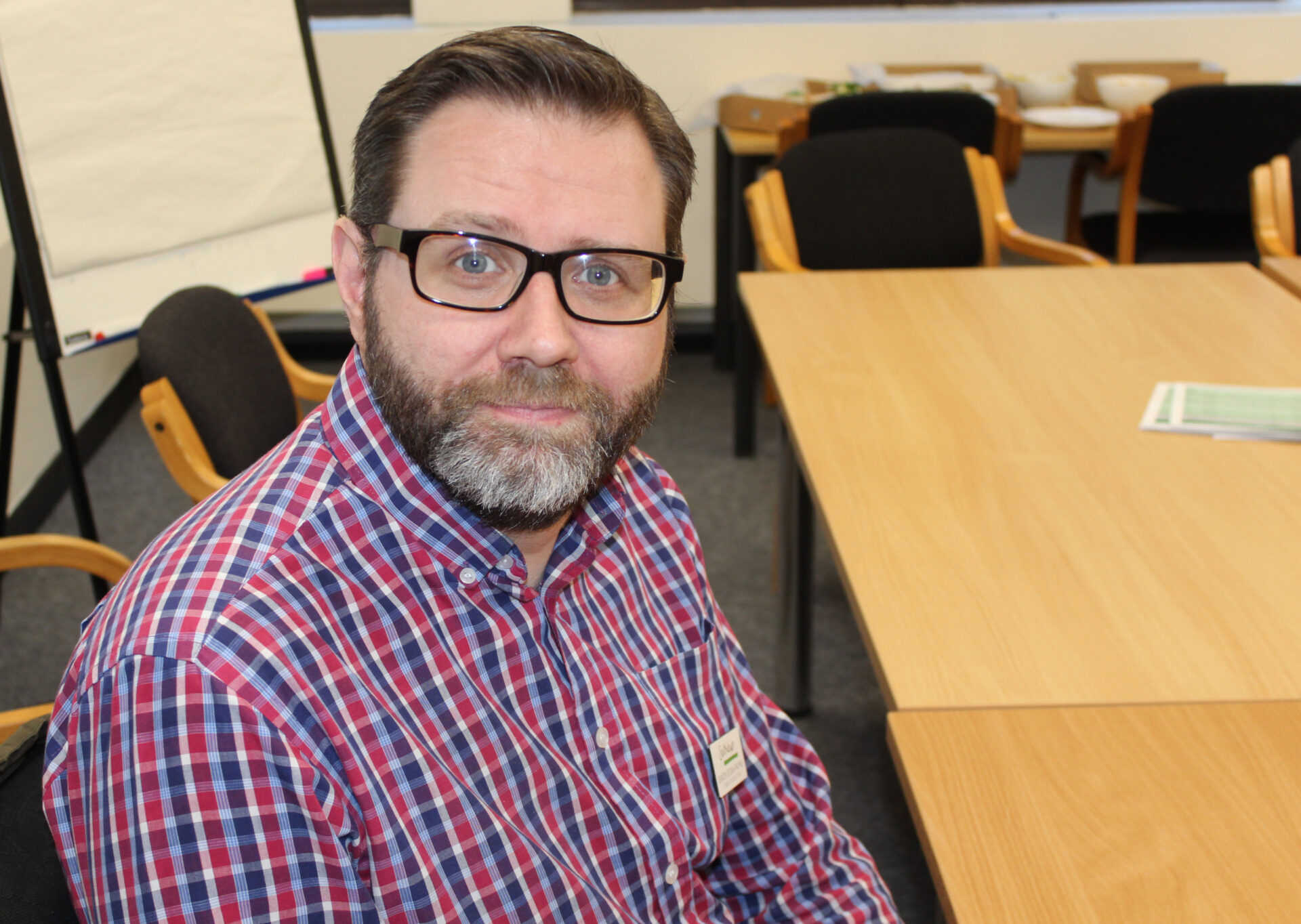 A man with brown hair and glasses and a checked shirt looks at camera