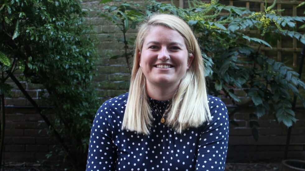 A woman with long blonde hair sits in a garden smiling at the camera