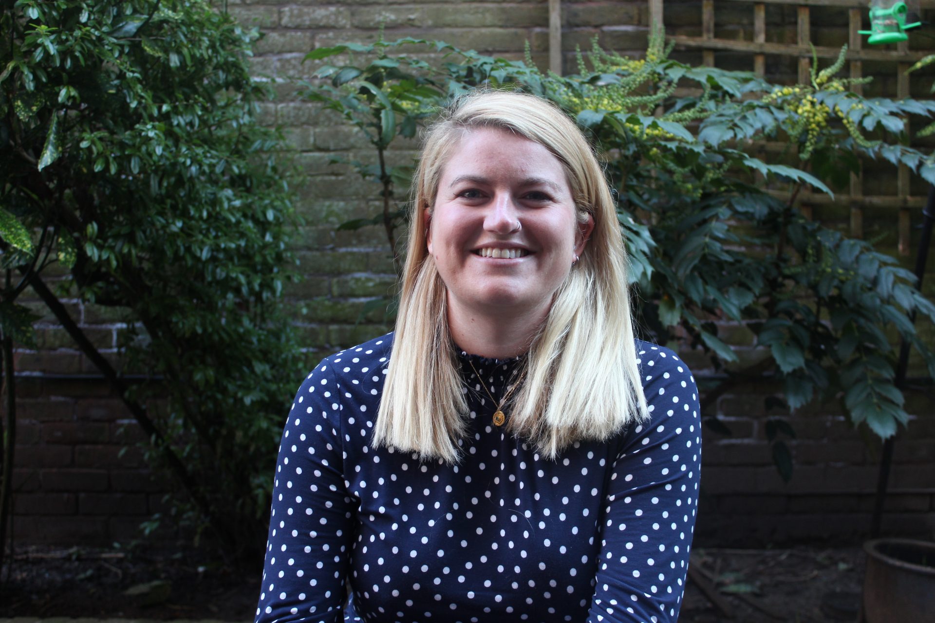 A woman with long blonde hair sits in a garden smiling at the camera