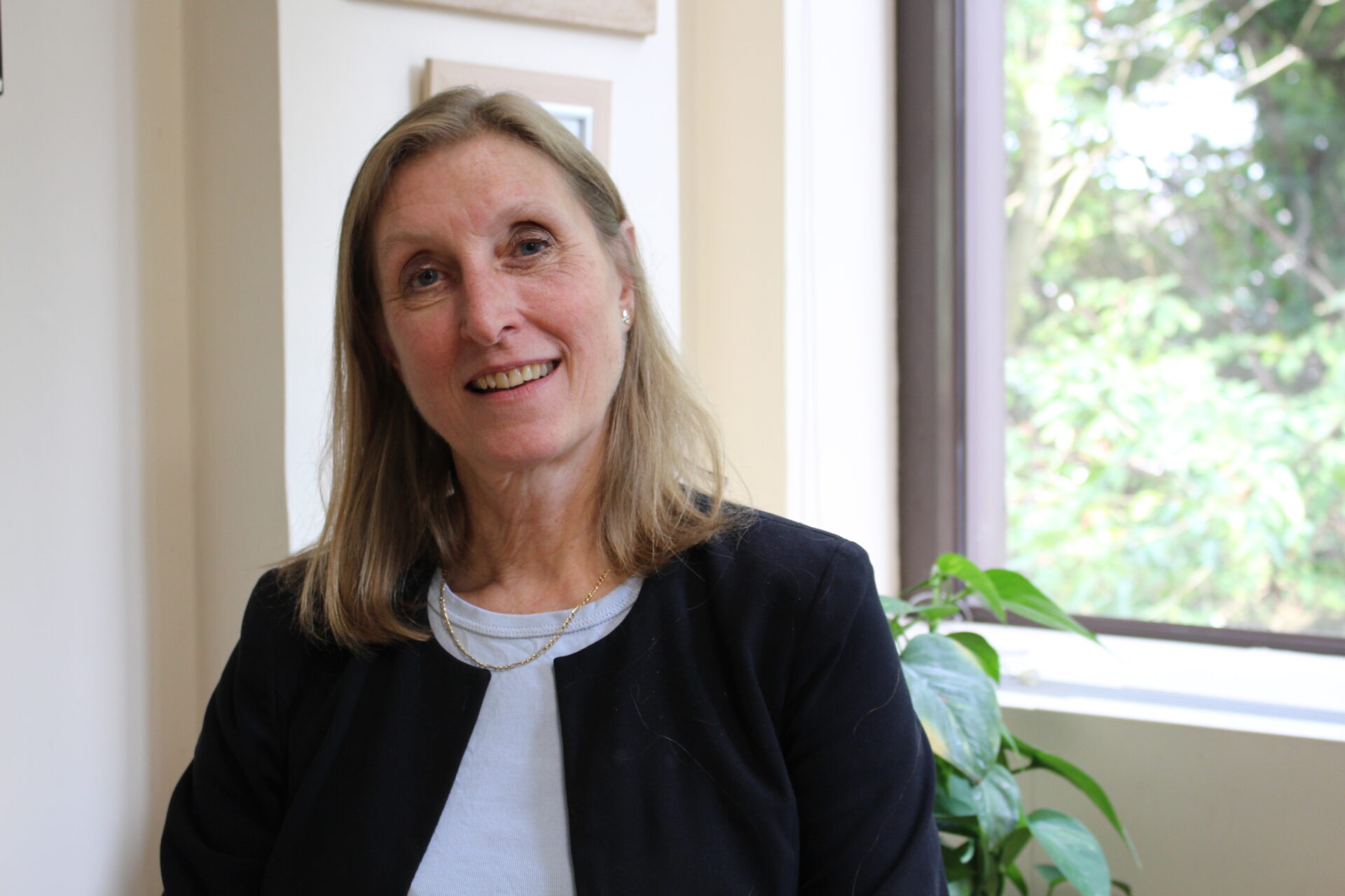 Jane wears mid-length blonde hair and a black jacket. There is a window behind her.