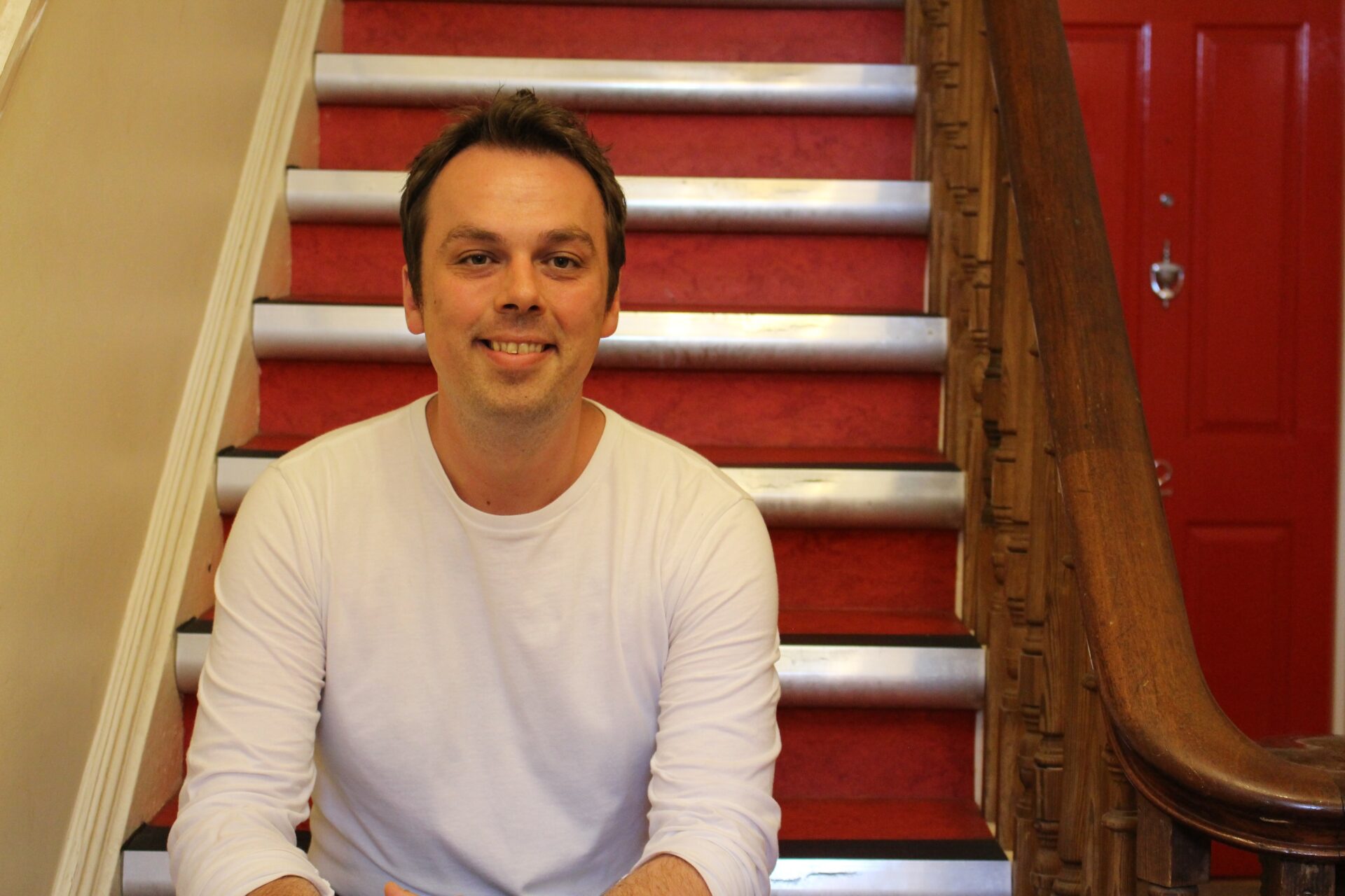 A man with short brain hair and a white t-shirt sits on some stairs and smiles at camera