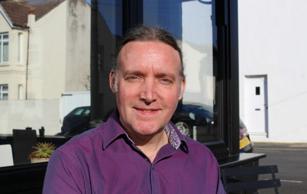 A man in a purple shirt sits outside a cafe looking at the camera