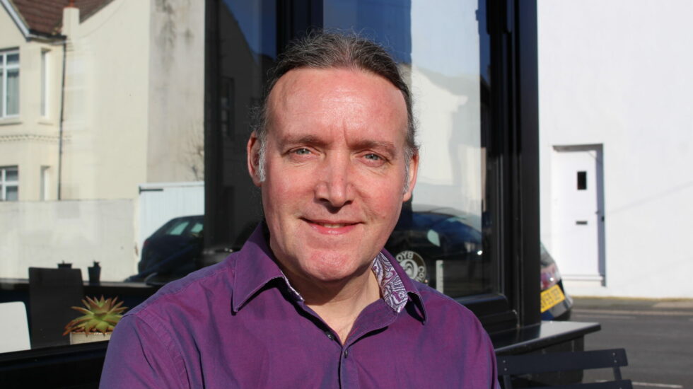 A man in a purple shirt sits outside a cafe looking at the camera