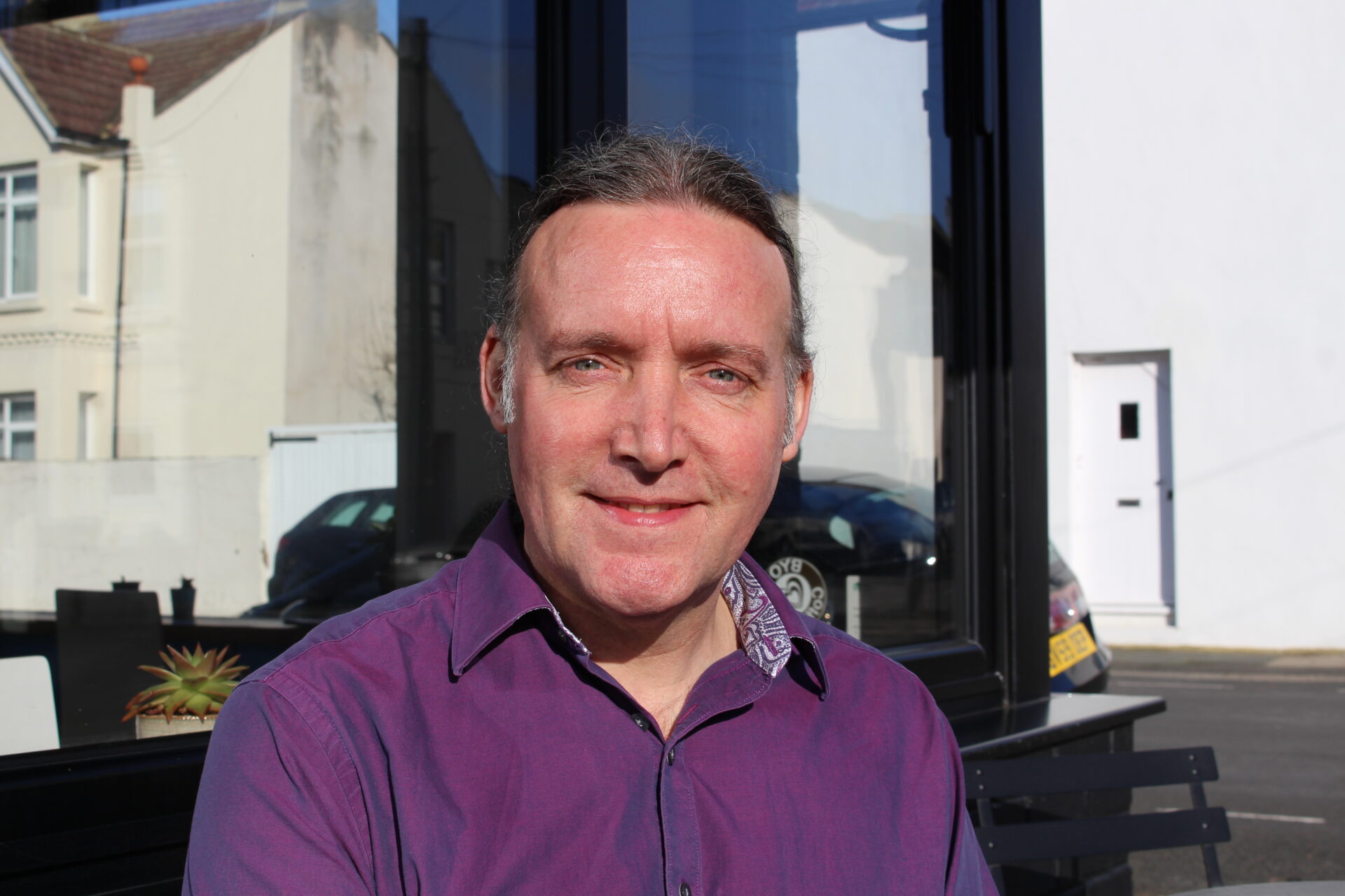 A man in a purple shirt sits outside a cafe looking at the camera