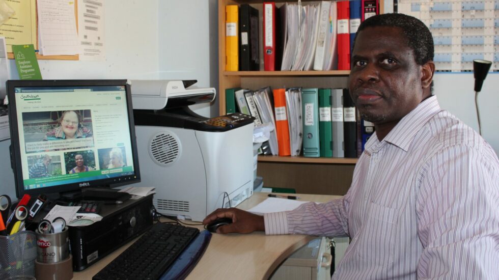 A black man with short black hair sits at his computer in his office and looks at the camera