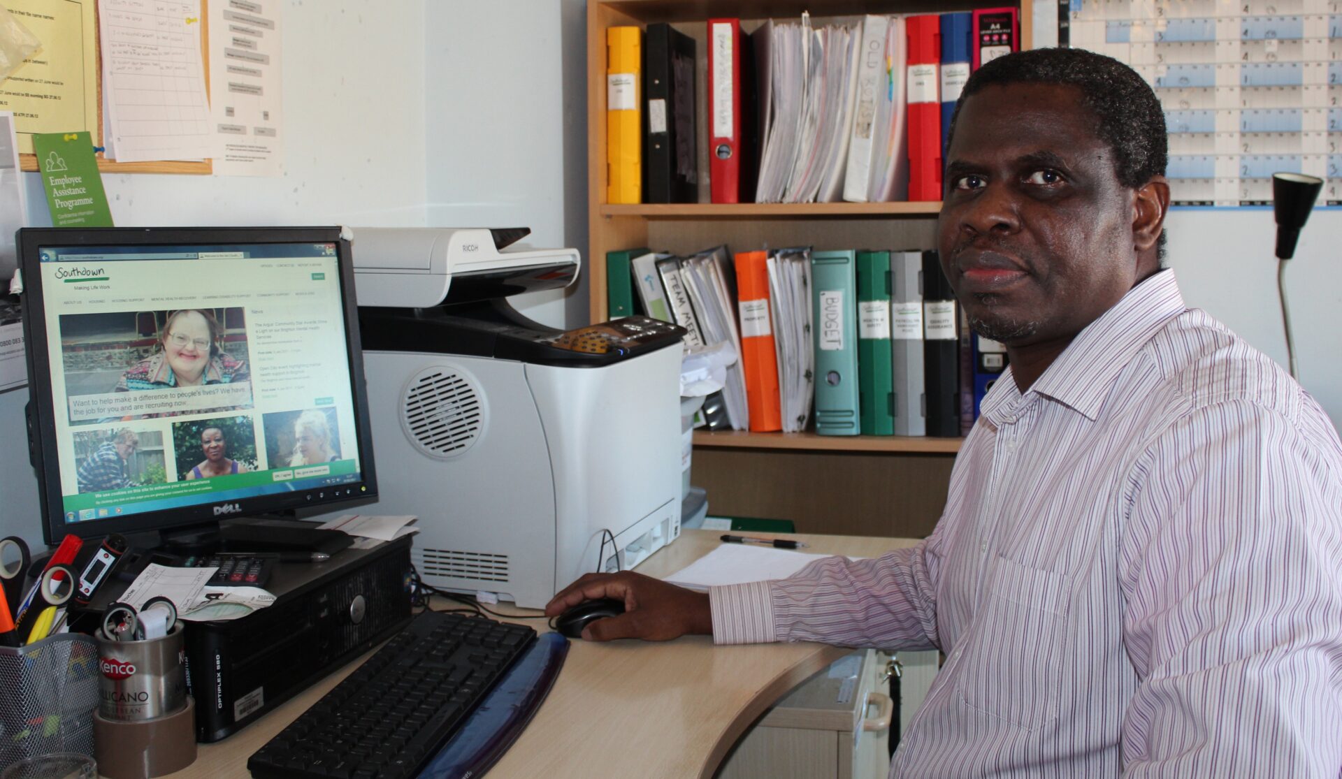 A black man with short black hair sits at his computer in his office and looks at the camera