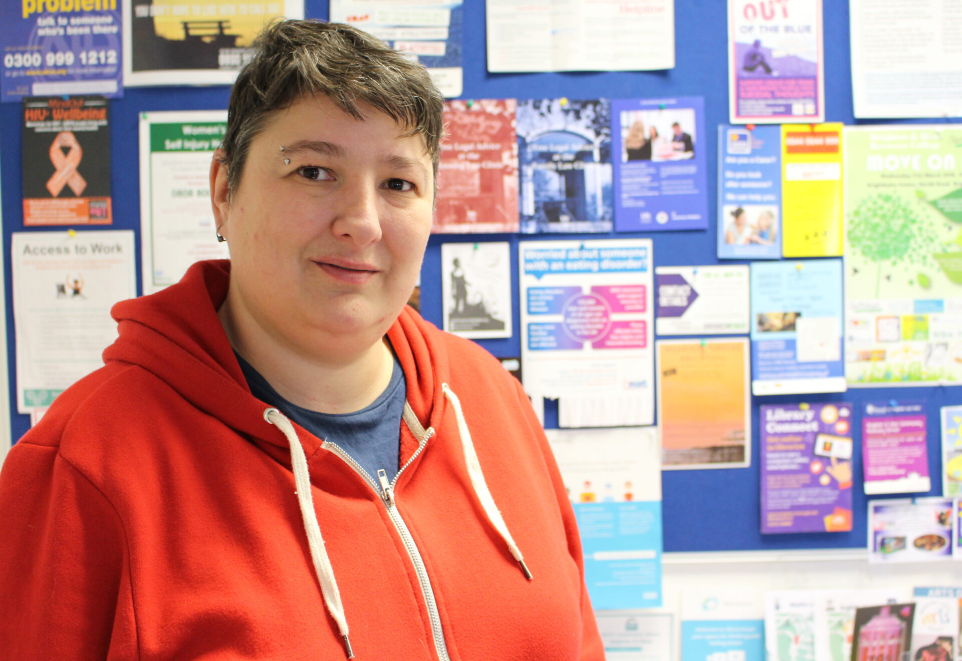 A person with short brown hair wears a bright orange hoodie and stands in front of a notice board