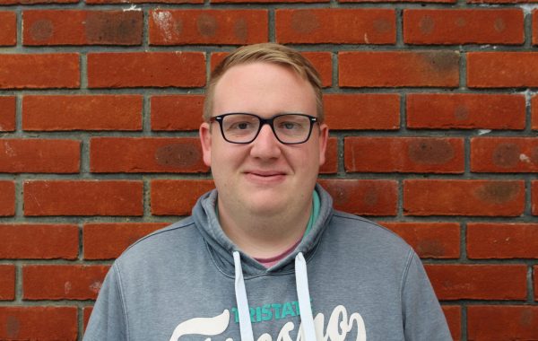 A man with short blond hair and dark glasses stands in front of a wall with a smile to the camera