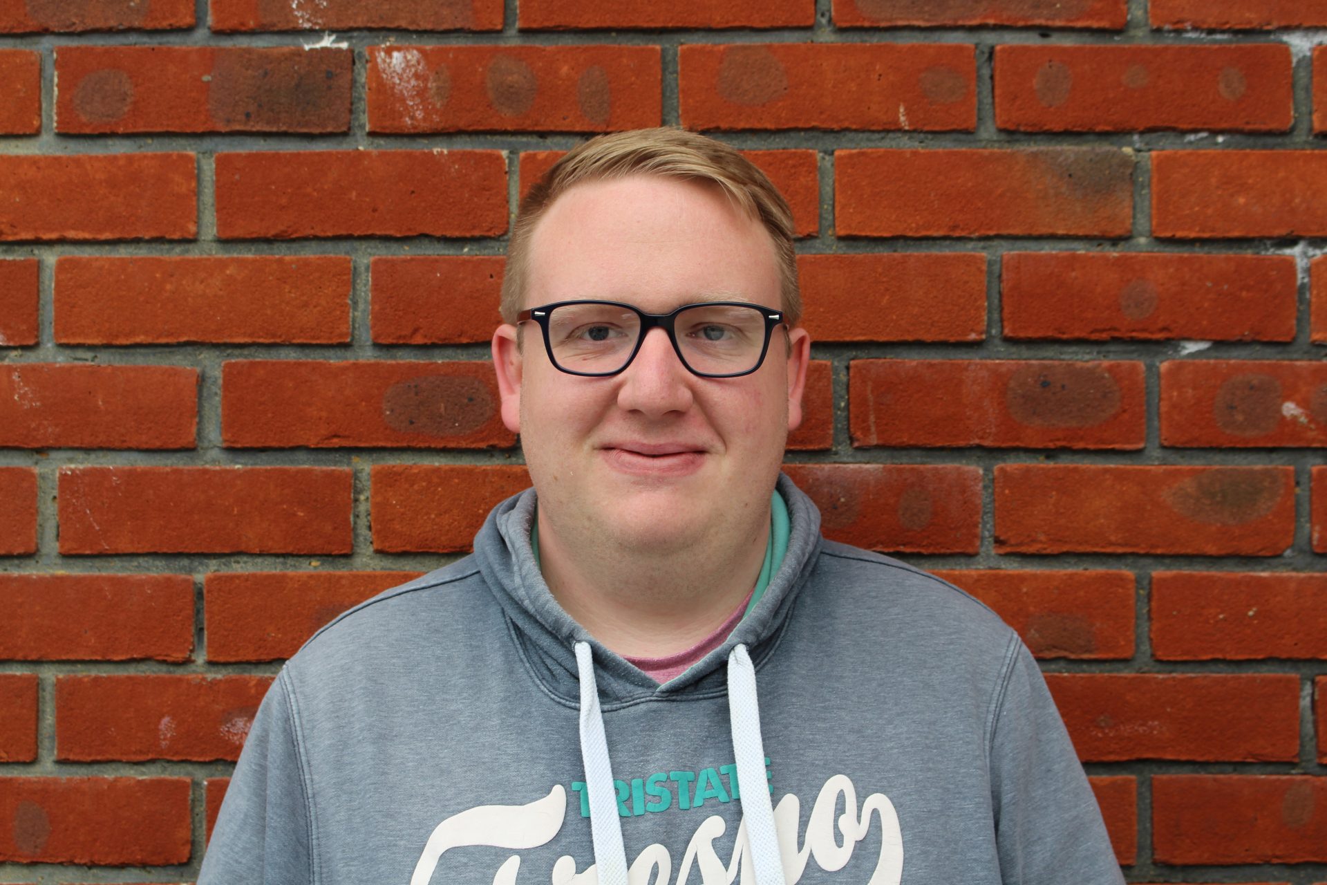 A man with short blond hair and dark glasses stands in front of a wall with a smile to the camera