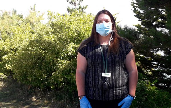 A woman stands outside wearing a mask and gloves, looking at the camera
