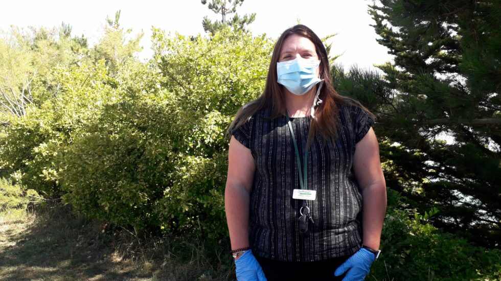 A woman stands outside wearing a mask and gloves, looking at the camera