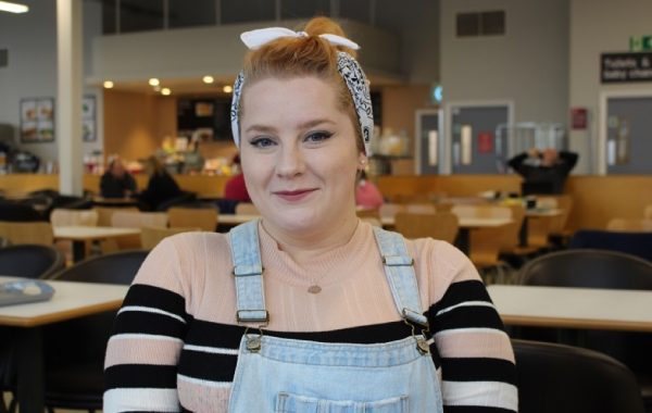 Close up photo of person smiling, wearing casual clothes with cafe seating in the background