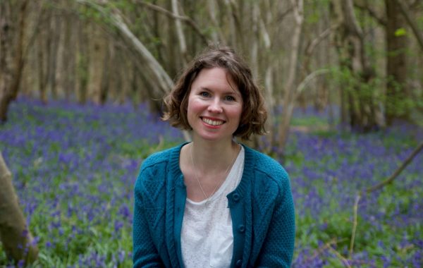 A woman with bobbed brown hair smiles at the camera. She is wearing a teal cardigan and white t-shirt and is surrounded by bluebells in a wood.