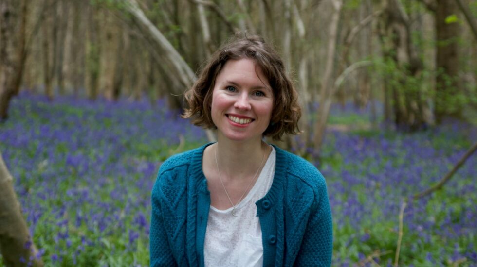 A woman with bobbed brown hair smiles at the camera. She is wearing a teal cardigan and white t-shirt and is surrounded by bluebells in a wood.