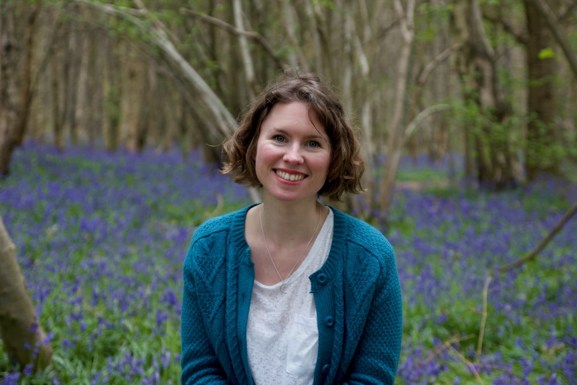 A woman with bobbed brown hair smiles at the camera. She is wearing a teal cardigan and white t-shirt and is surrounded by bluebells in a wood.