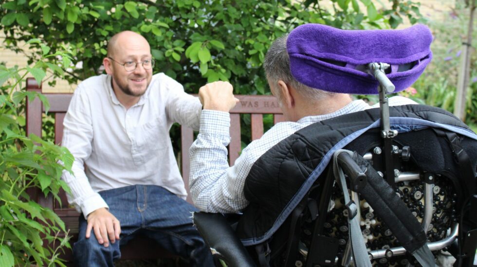 a man wearing glasses and a shirt hands some fruit to a client in a wheelchair in the garden