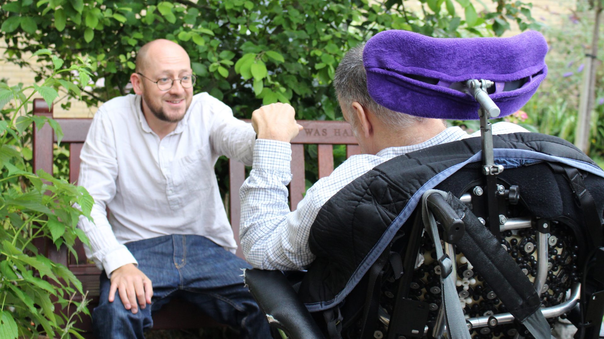 a man wearing glasses and a shirt hands some fruit to a client in a wheelchair in the garden