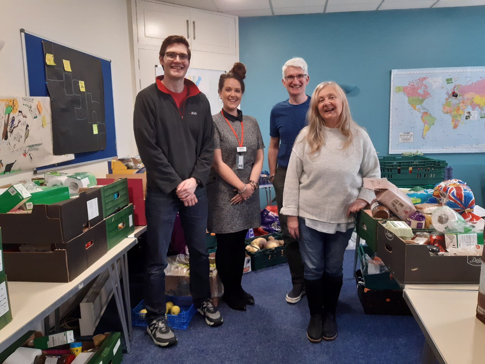 Four individuals are standing in a room looking at the camera and smiling. The room is filled with boxes of donated food.