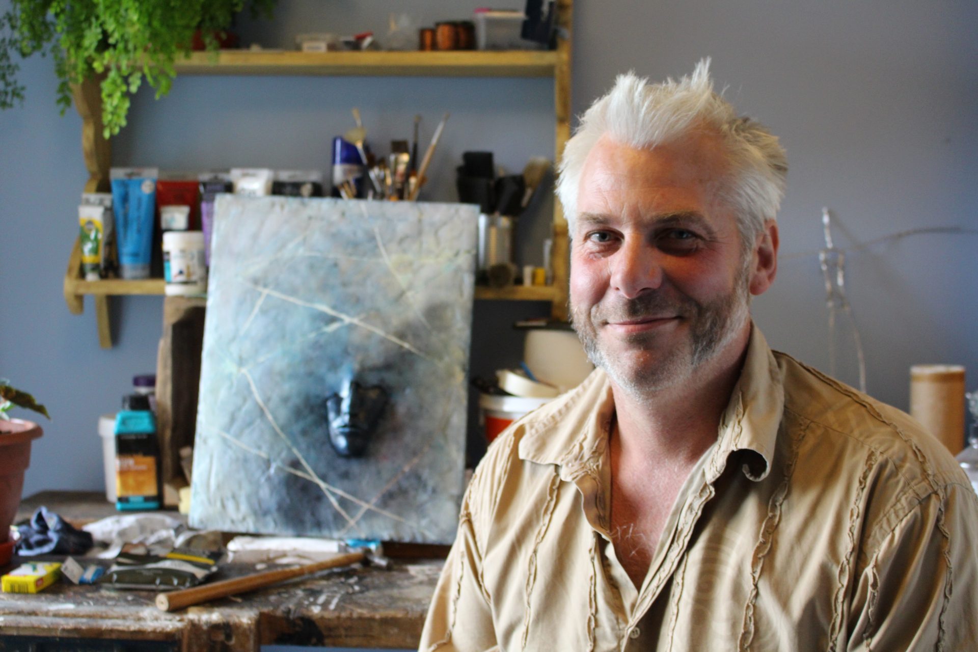 A man sits smiling at the camera. Behind him is a self-portrait he created.