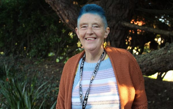 A Support Worker with short blue hair smiles at the camera. She is wearing a dark orange cardigan, stripey top, and a lanyard around her neck.