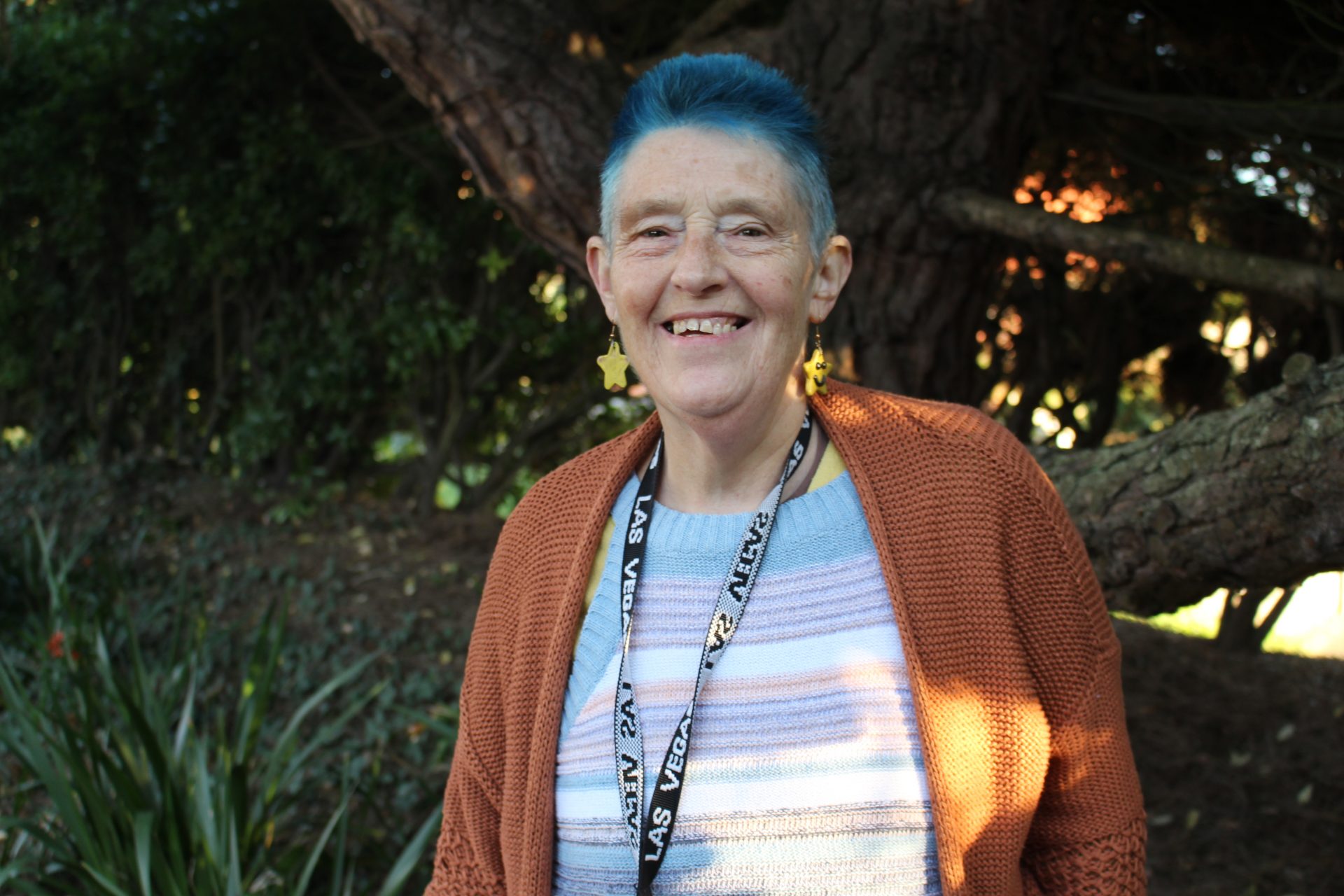 A Support Worker with short blue hair smiles at the camera. She is wearing a dark orange cardigan, stripey top, and a lanyard around her neck.