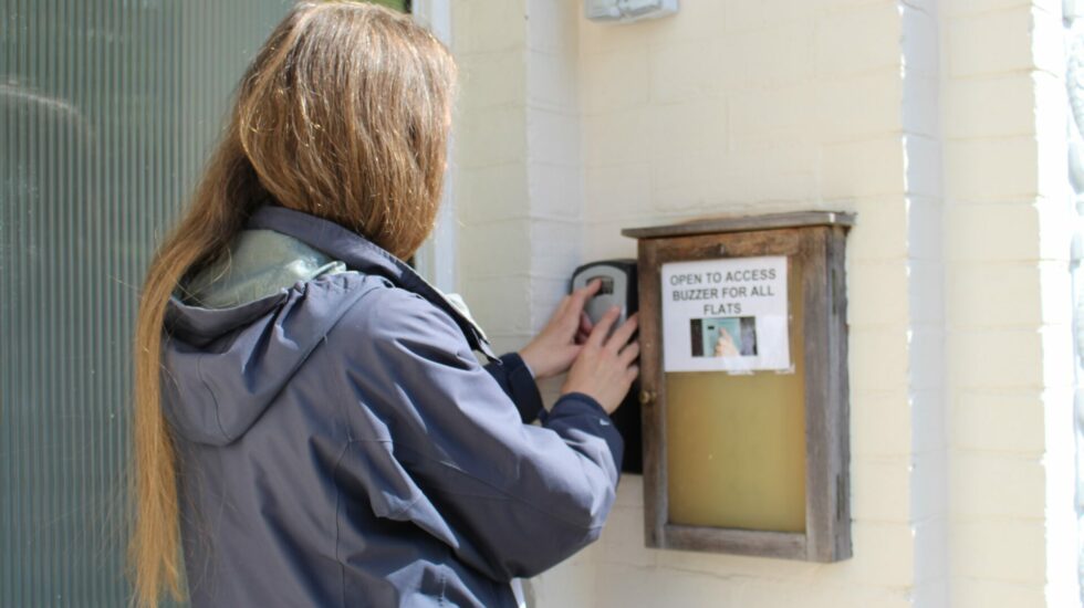 A woman with long hair wearing a raincoat stands outside a house and presses the buzzer