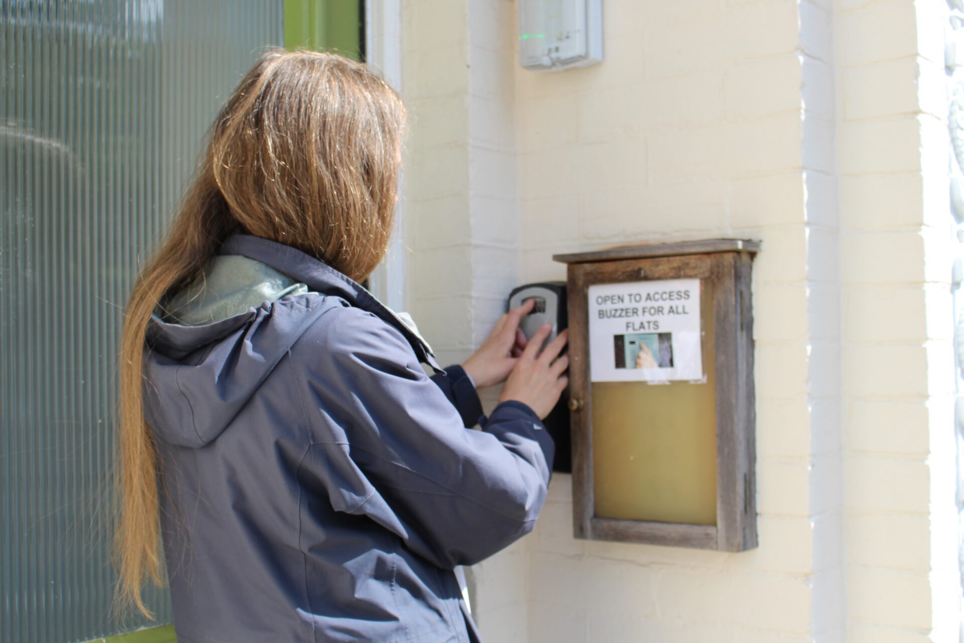A woman with long hair wearing a raincoat stands outside a house and presses the buzzer