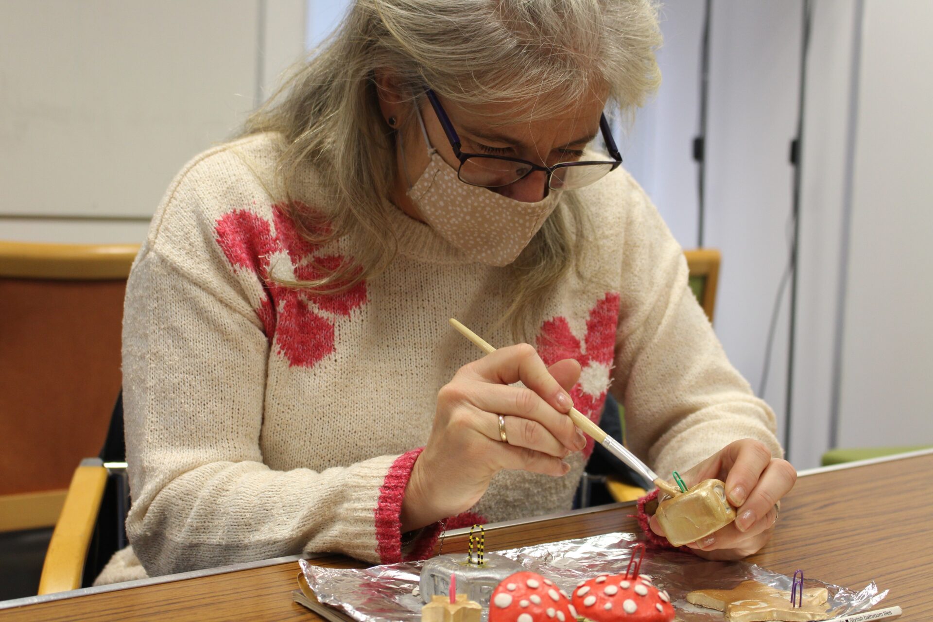 A woman with mid-length hair wearing glasses and a mask paints an object