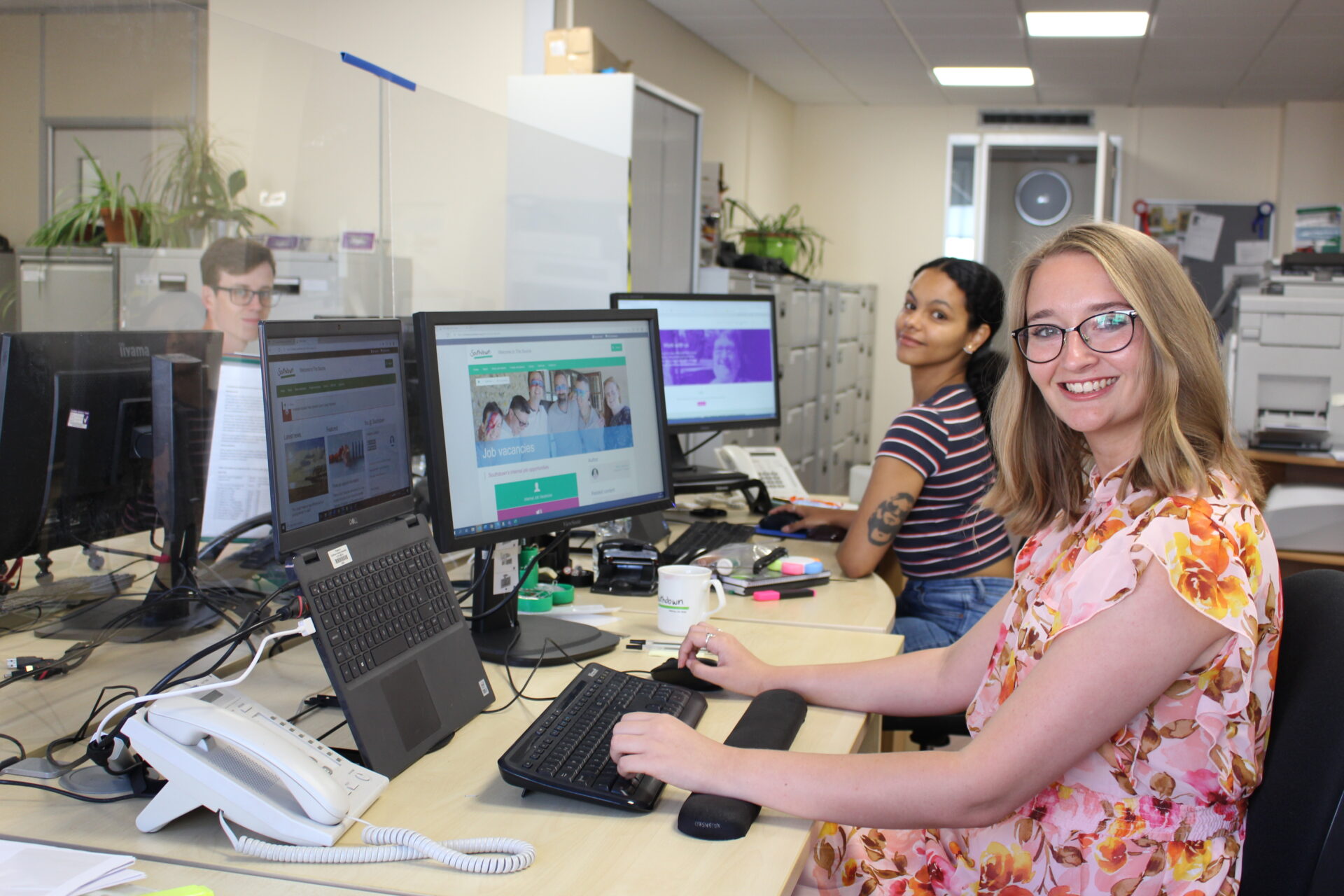 A woman with blonde hair and glasses smiles widely at the camera. Her two colleagues sit at their computers behind her. They are in an office.