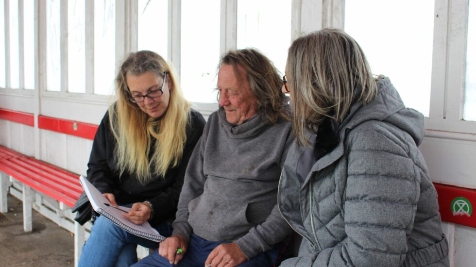 Photo of two Support Officers sitting either side of a client in a bus shelter looking at paperwork together