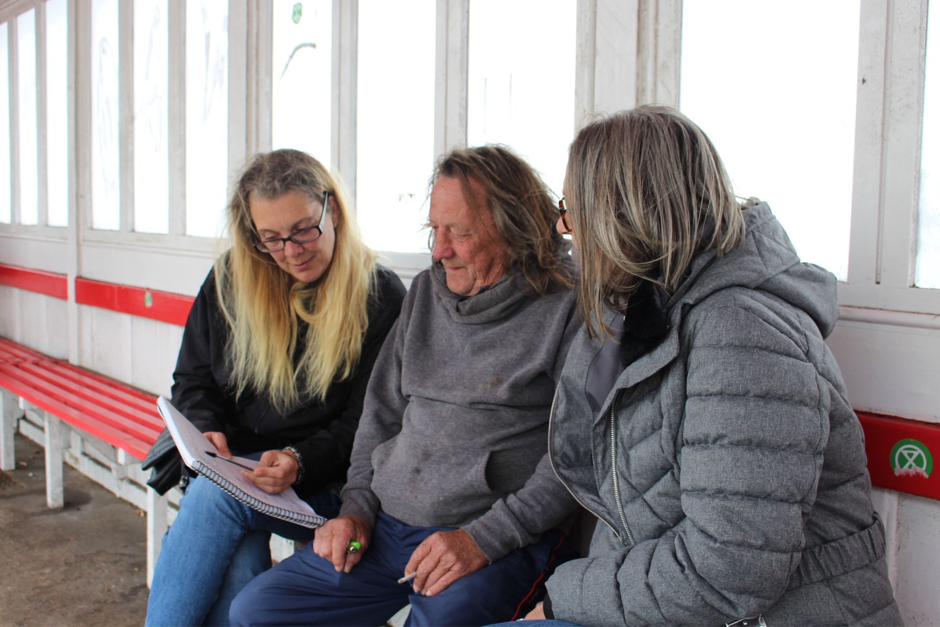 Photo of two Support Officers sitting either side of a client in a bus shelter looking at paperwork together