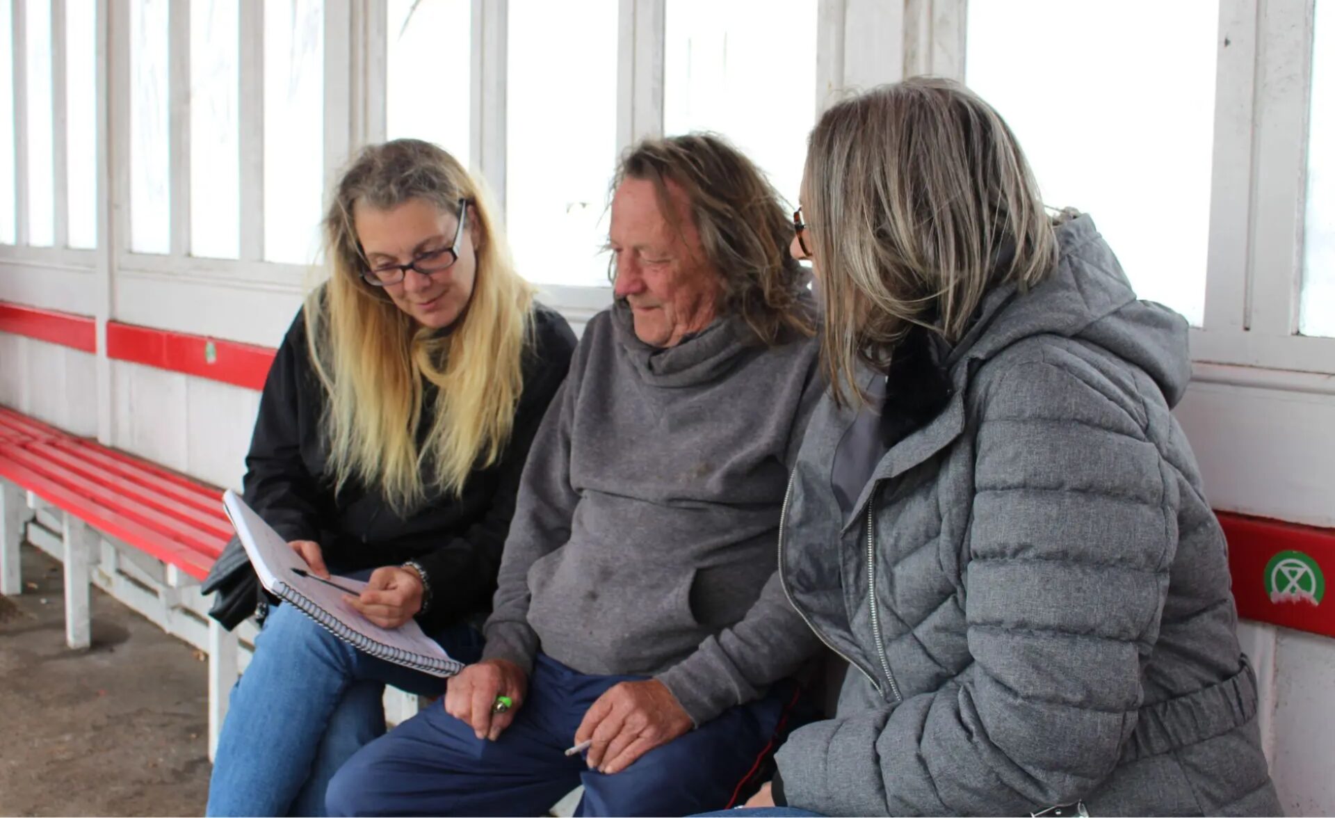 Photo of client sitting in bus shelter side by side between two workers, looking at a form together