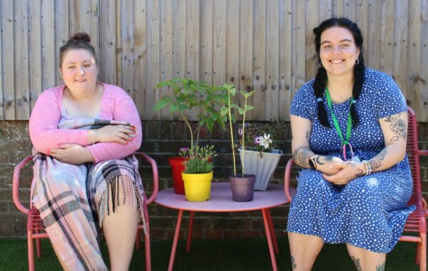 A client wears a pink cardigan and she is hugging a blanket. She sits next to a Support Worker in blue dress and dark pigtails.