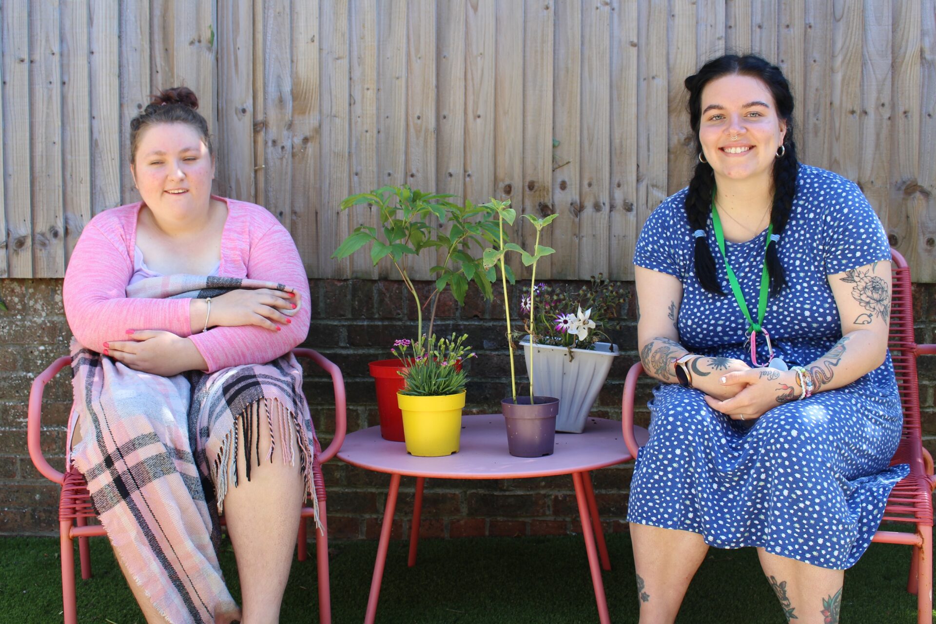 A client wears a pink cardigan and she is hugging a blanket. She sits next to a Support Worker in blue dress and dark pigtails.