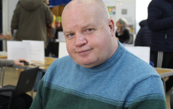 A white man with fair skin looks directly at the camera with a smile. He is wearing a blue and green jumper.