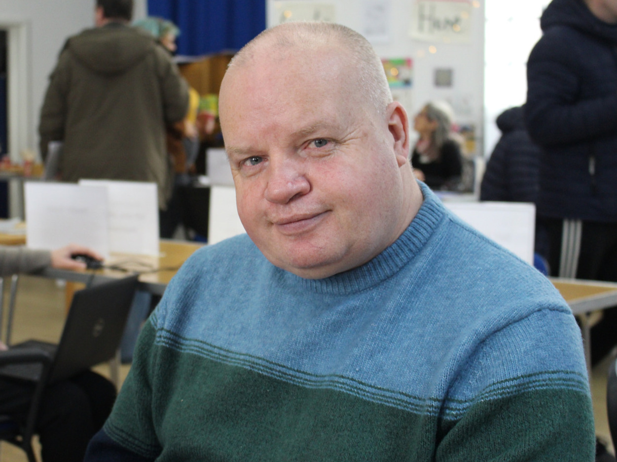 A white man with fair skin looks directly at the camera with a smile. He is wearing a blue and green jumper.