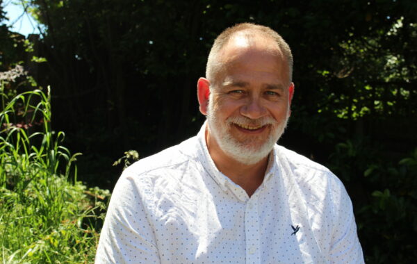 A man with a white beard and white shirt sits in the sun looking directly at the camera