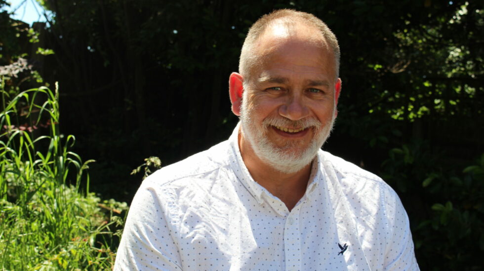 A man with a white beard and white shirt sits in the sun looking directly at the camera