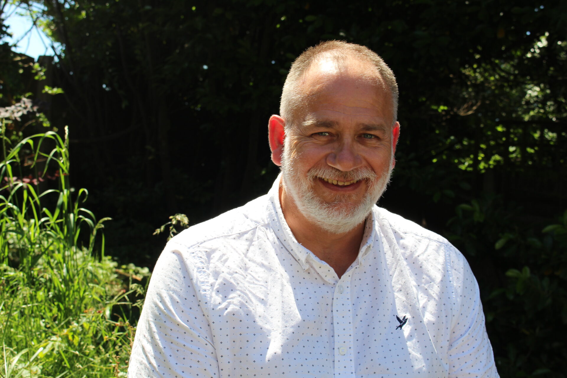 A man with a white beard and white shirt sits in the sun looking directly at the camera