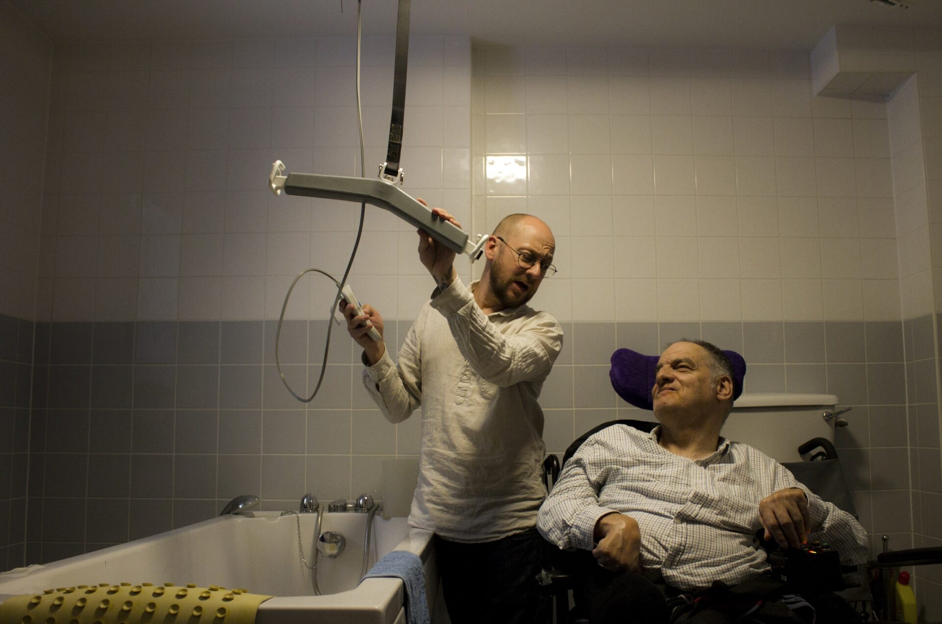 A white man is holding adapted bathroom equipment whilst a white client in a wheelchair watches him. The lighting is low and atmospheric.