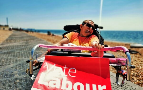 A white man in his wheelchair is campaigning for his political party on the beach in full sunshine.