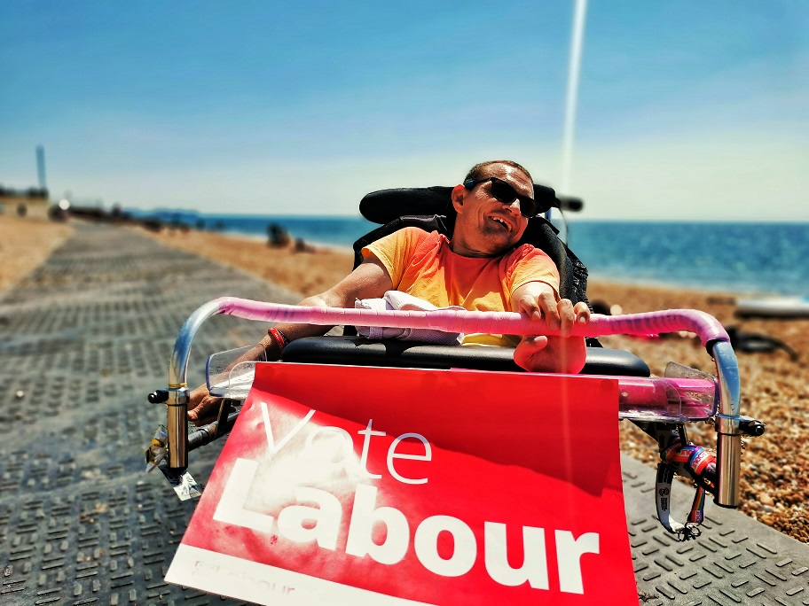 A white man in his wheelchair is campaigning for his political party on the beach in full sunshine.