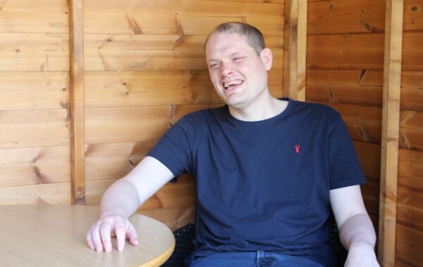 A white man wearing a black t-shirt sits in his summer house with a huge smile on his face.